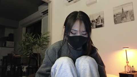 Media: Video of an Asian woman with long black hair, wearing a gray sweater, white pants, and a black face mask, sitting on a couch, in a dimly lit living room with a plant, wall art, and a lamp.