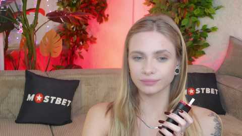 Media: Video of a blonde woman with tattoos, holding a vape pen, seated on a beige couch with black \"MOSTBET\" pillows, surrounded by green plants and red lighting.