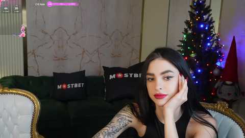 Media: Video of a woman with long black hair, red lipstick, and tattoos, sitting on a dark green couch with black pillows, in a room with a lit Christmas tree and colorful lights, wearing a sleeveless black top.