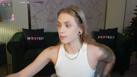 Media: Video of a young, light-skinned woman with blonde hair, tattoos, and black ear gauges, wearing a white tank top, sitting indoors with green couches and \"MOSTBET\" pillows in the background.