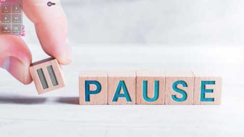 Media: Video: A hand holds a wooden block with \"Pause\" written in blue letters, placed on a white, tiled floor. The background is softly blurred.