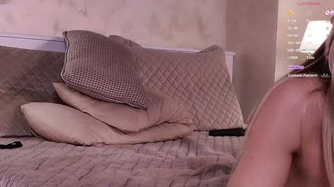 Media: Video of a woman with light skin, lying on a bed with beige quilted bedding and pillows, partially covered by a checkered blanket. The room has beige walls, and a video game interface with player stats and achievements is visible on the right.