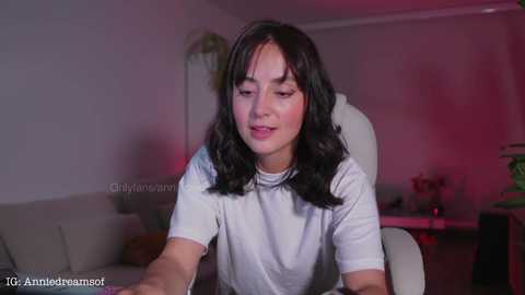 Media: A video of a young woman with long, dark hair, wearing a white top, sitting on a white chair in a dimly lit, modern living room with pink lighting.