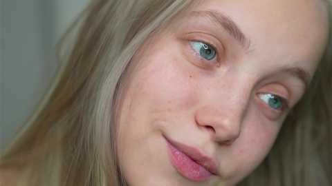 Media: Video of a close-up of a young Caucasian woman with fair skin, blue eyes, and blonde hair. She has a slight smile, freckles, and a soft, natural makeup look. The background is blurred and light-colored.