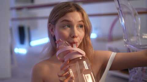 Media: Video of a blonde woman with fair skin, wearing a white tank top, drinking from a pink sippy cup, in a modern kitchen with blue and pink lighting.