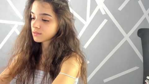 Media: Video of a young girl with long, wavy brown hair, light skin, and a small build, wearing a white strapless dress. She is in a modern, minimalist room with white geometric patterns on the walls and a black office chair visible in the background.