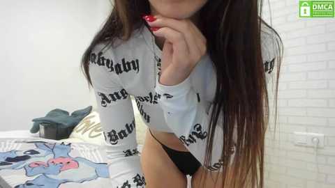 Media: A video of a young woman with long brown hair, wearing a white \"baby\" sweatshirt and black panties, standing in a minimalist bedroom with white brick wall and a bed with a blue comforter.