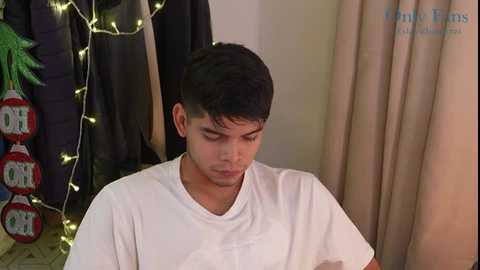 Media: Video of a young man with short dark hair wearing a white t-shirt, looking down, in a room with beige curtains, a green plant, and hanging string lights.