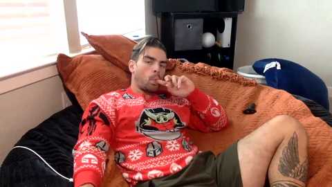 Media: Video of a man in a red Star Wars-themed sweater, brown shorts, and a tattoo on his leg, lounging on an orange bedspread in a cozy, cluttered room with a black TV, blue chair, and orange pillows.