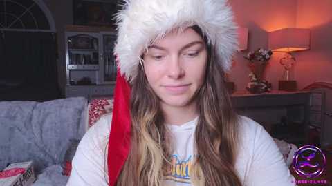 Media: A video of a young woman with long, brown hair wearing a fluffy white Santa hat with a red visor, a white T-shirt, and a neutral expression, set in a cozy, warmly lit living room.