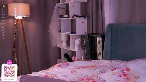 Media: A video of a cozy, modern bedroom with a white bed covered in a pink floral duvet, a white shelving unit, and a tall floor lamp.