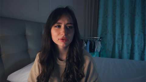 Media: Video of a young woman with long dark hair, wearing a beige top, looking contemplative in a dimly lit bedroom with white sheets, a gray headboard, and teal curtains.