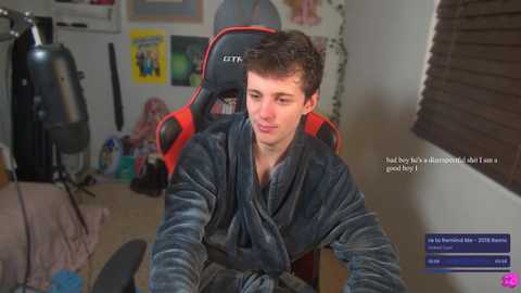 Media: Video of a young white man with short brown hair, wearing a plush robe, sitting in a red gaming chair in a cluttered room.