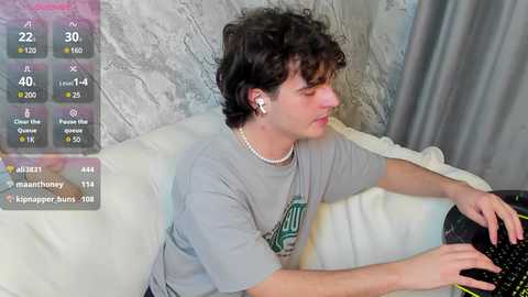 Media: Video of a young man with curly hair, wearing a grey t-shirt, playing a video game on a black controller, in a cozy living room with a textured grey wall.