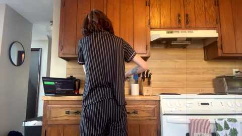 Media: A video of a person with shoulder-length brown hair, wearing a black and white striped jumpsuit, cooking in a modern kitchen with wooden cabinets, white appliances, and a tiled backsplash.