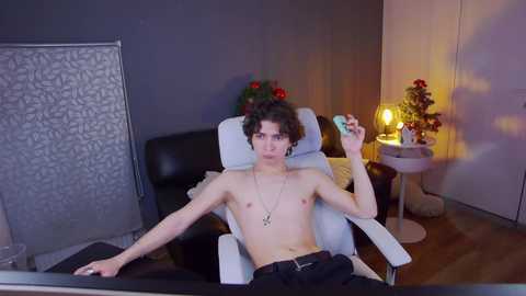 Media: Video of a shirtless, slim, curly-haired man with fair skin reclining in a black leather chair, smoking a cigarette in a dimly-lit, cozy room with a cat, a small table, and festive decorations.