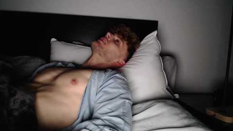 Media: Video of a shirtless, fit, young man with short brown hair lying on a bed with white pillows, wearing an open light blue shirt, in dim lighting.