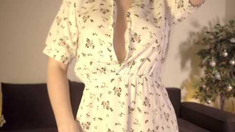 Media: Video of a woman wearing a white, short-sleeved dress with a floral pattern, featuring a deep V-neckline. The background includes a dark brown couch and a potted plant.