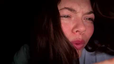 Media: Close-up video of a woman with long, dark hair, brown eyes, and light skin, puckering her lips in a suggestive kiss. Background is dark and indistinct.