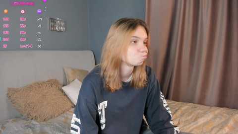 Media: A video of a young woman with shoulder-length blonde hair, wearing a dark blue sweatshirt, sitting on a bed in a dimly lit room with a TV displaying a digital channel guide.