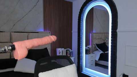 Media: Video of a modern bedroom with a large, illuminated, arched mirror on the right. A life-sized, realistic dildo is inserted into a man's hand, which is resting on a black bed with white pillows and a beige headboard. The background features striped wallpaper and a white door.