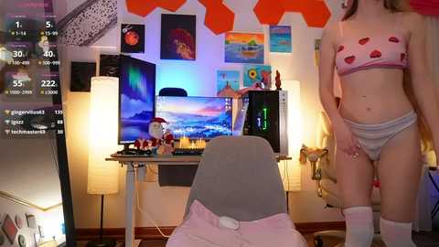 Media: Video of a young, slender, light-skinned woman in a pink heart-patterned bra and panties, standing in a brightly lit, colorful room with a computer desk, monitors, and wall art.