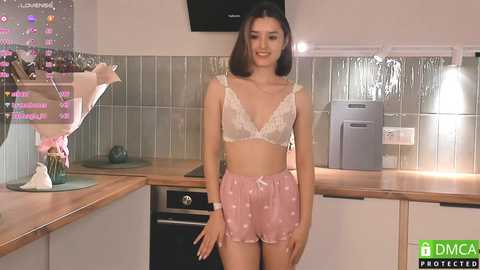 Media: Video of a young woman with light skin and straight black hair in a beige lace bra and pink shorts, standing in a modern kitchen with tiled backsplash and stainless steel appliances.