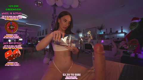 Media: A video featuring a slender, light-skinned woman with long brown hair in a white bikini, pouring a clear liquid onto a large, erect penis in a dimly-lit, festive room.