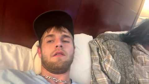 Media: Video of a young man with short brown hair, beard, wearing a black cap and chain necklace, lying on a bed with white and patterned sheets against a dark wooden headboard.