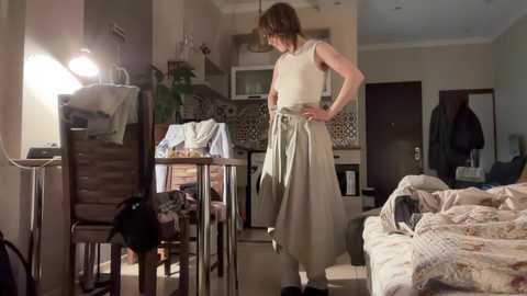 Media: Video of a woman in a white sleeveless top and beige skirt, standing in a dimly lit, messy kitchen.