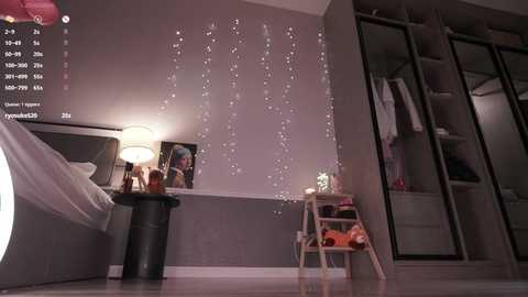 Media: A video of a modern bedroom with a gray wall adorned with fairy lights, a bed with white sheets, a black stool, and a wooden chair with a colorful toy box.