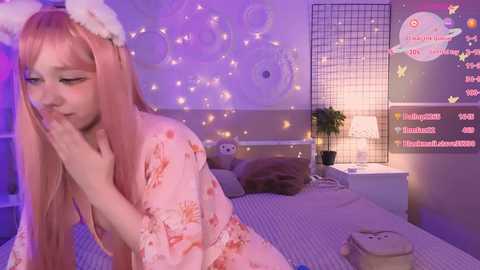 Media: A video of a young woman with pink hair, wearing a floral robe, sitting on a bed with a white lamp, and a chat box on a wall in a purple-lit room.