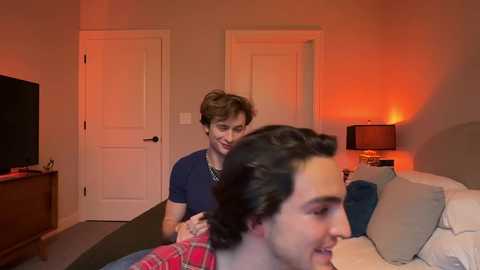 Media: Video of a cozy bedroom with two people, one with curly brown hair in a dark shirt, and another with dark hair in a plaid shirt, smiling. Red lights cast a warm glow.