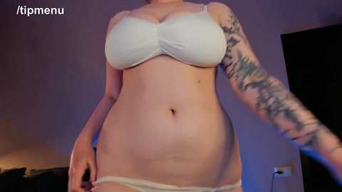 Media: Video of a light-skinned woman with large breasts and a tattooed arm, wearing a white bra and panties, standing indoors.