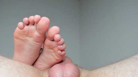 Media: Video of a man's bare feet and legs against a plain, light gray background. His feet are relaxed, with visible soles and toenails. The skin appears smooth and lightly tanned. The focus is on the feet, with the legs extending out of frame.