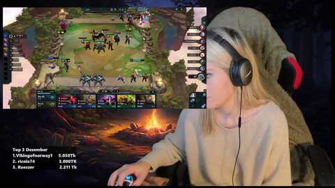 Media: Video of a young girl playing League of Legends on a computer, wearing a beige hoodie and headphones, set against a dark room with a campfire and rugged landscape in the background.