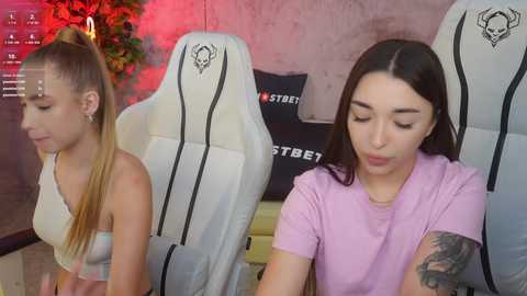 Media: Video of two young women, one with long blonde hair in a white top, the other with long dark hair in a purple shirt, sitting on white gaming chairs, background includes a red flower arrangement and gaming-themed decor.