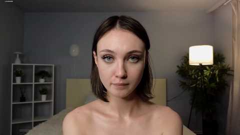 Media: Video of a young woman with fair skin, shoulder-length brown hair, and blue eyes, standing in a dimly-lit, minimalist bedroom with grey walls, a white shelf, a potted plant, and a tall lamp.