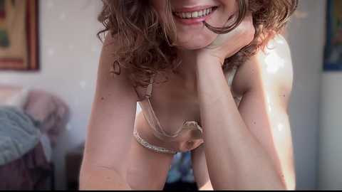 Media: A video of a smiling woman with curly brown hair, wearing a beige bra, leaning forward, with a blurred background featuring a framed painting and a bed.