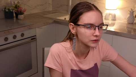 Media: Video of a young woman with glasses and long brown hair, wearing a pink t-shirt and apron, seated in a modern kitchen with white cabinets and marble countertops.