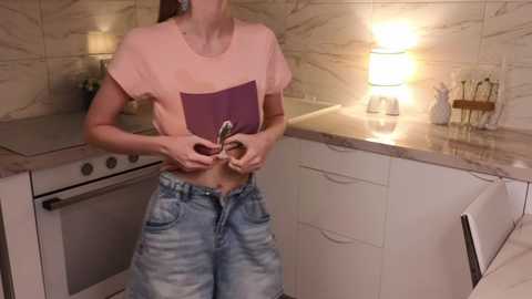 Media: Video of a woman in a modern kitchen, lifting her pink t-shirt to reveal a purple pouch, wearing light blue jeans.