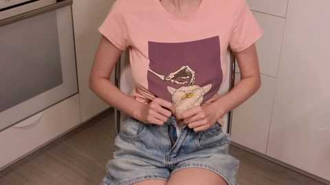 Media: Video of a young person with light skin, sitting in a kitchen, wearing a pink t-shirt with a white and purple graphic design, and light blue denim shorts. The person holds a plush toy resembling a skeleton.