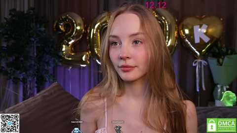 Media: Video of a young blonde woman with fair skin, wearing a pink top, standing in a room with large golden \"2019\" balloons and a plant.