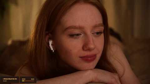 Media: Video of a young woman with fair skin and long red hair, listening to headphones, lying on a bed, looking contemplative.