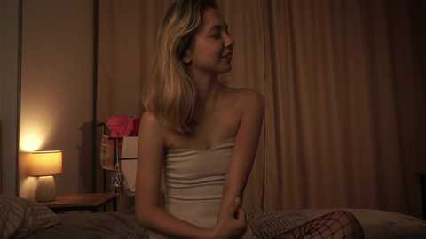 Media: A video of a blonde woman in a strapless beige dress, sitting on a bed, with dimly lit room in the background.