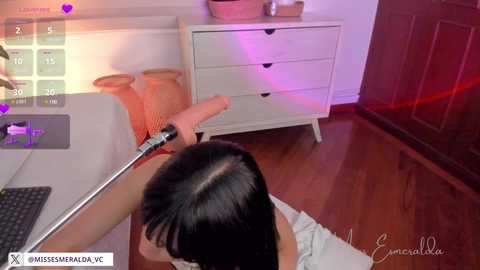 Media: A video shows a woman with dark hair kneeling on a polished wooden floor, aiming a dildo at a white dresser with drawers, under a pink light, in a modern room.