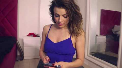 Media: Video of a woman with wavy brown hair, wearing a blue tank top, standing in a bedroom with red walls and a white door. She is focused on her phone.