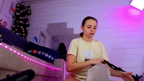 Media: A video shows a young woman with light skin and brown hair in a ponytail, wearing a beige T-shirt, sitting on a couch, holding a black device, with a Christmas tree and purple LED lights in the background.