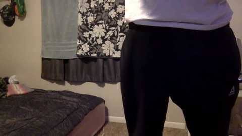 Media: A video of a person wearing black athletic pants and a light purple shirt standing in a room with a bed covered in dark bedding, a floral-patterned curtain, and a plant on the left.