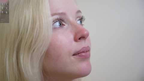 Video of a close-up side profile of a Caucasian woman with fair skin, light blonde hair, and blue eyes. She has natural makeup and a neutral expression. The background is a plain, light-colored wall.